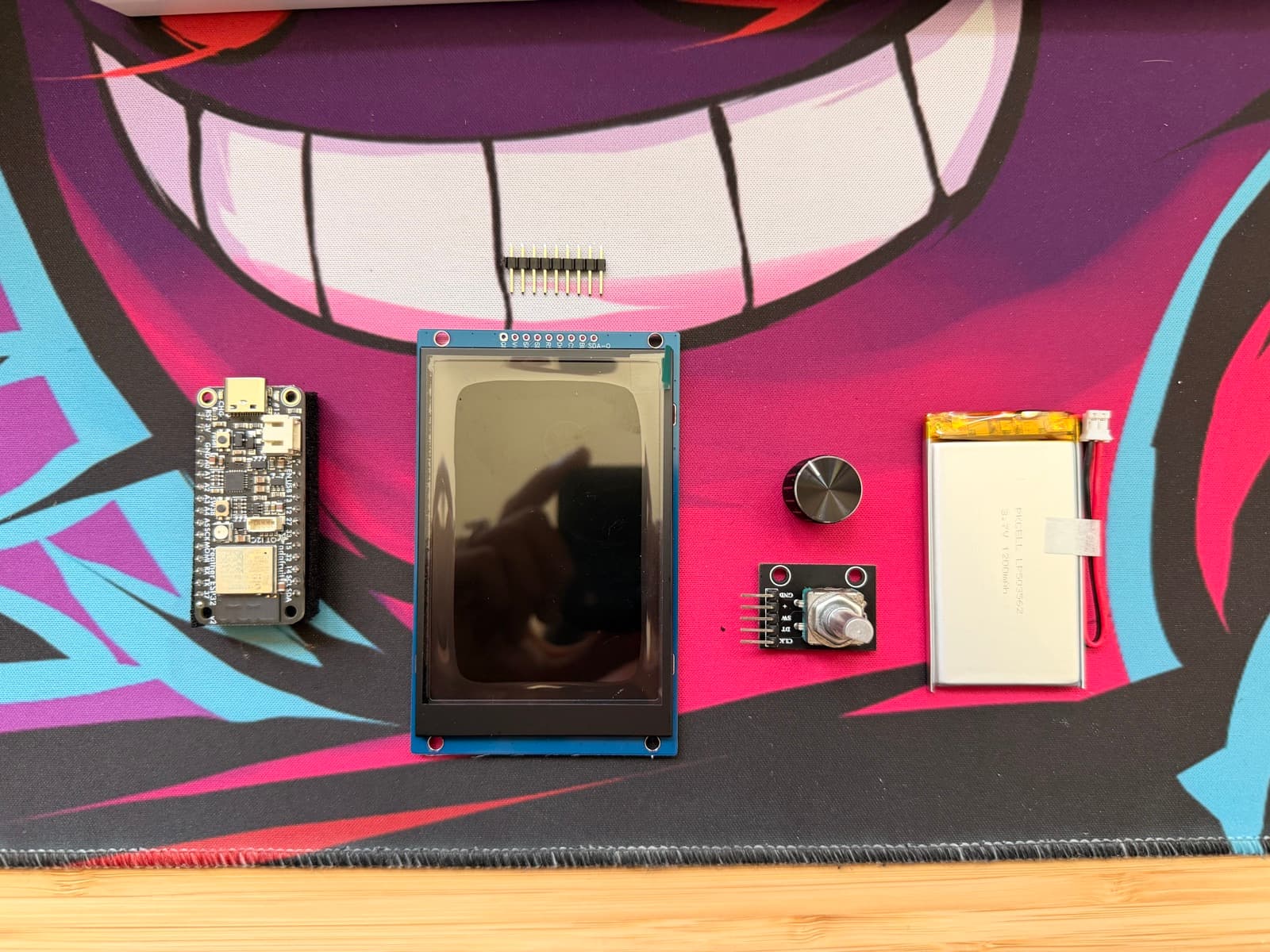 The e-reader parts laid out on a Gengar desk mat: the ESP32 Feather V2 board, the 3.5-inch TFT display, a rotary encoder with its knob, the 3.7V 1200mAh battery, and a strip of pin headers