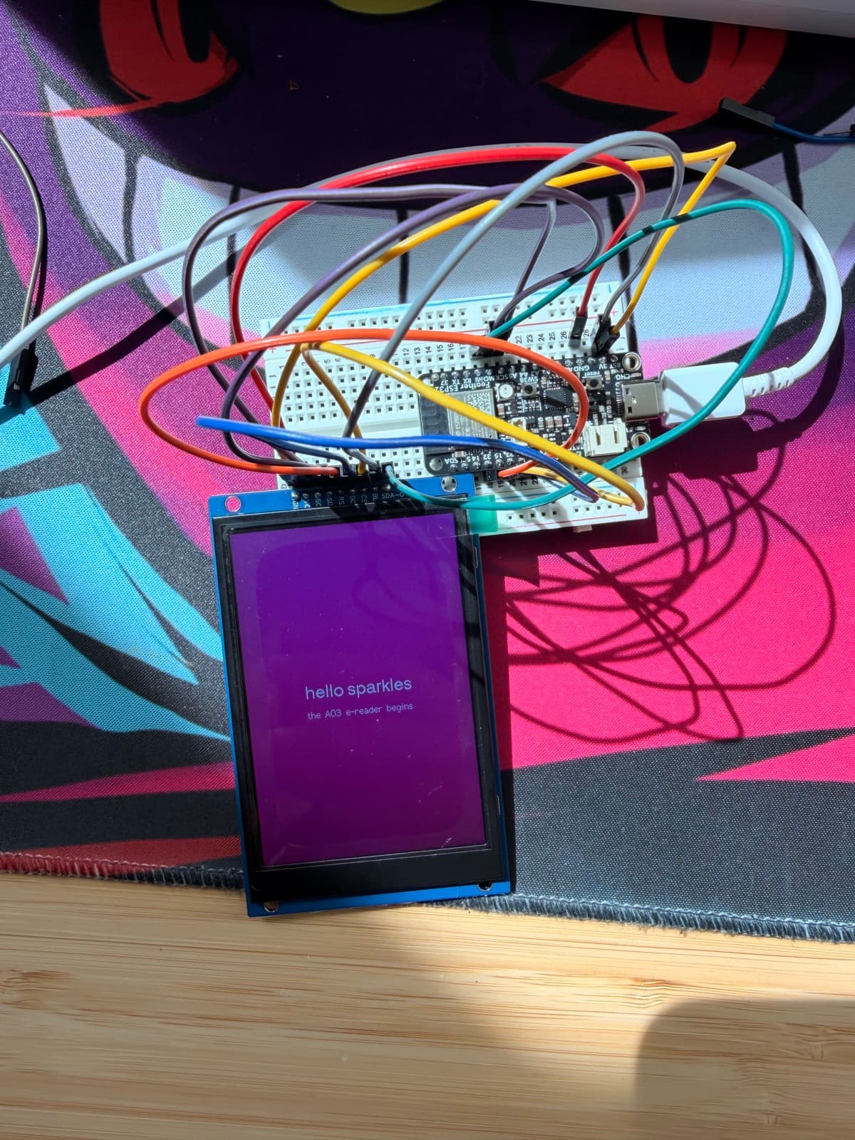 The 3.5-inch display rendering 'hello sparkles — the AO3 e-reader begins' in white text on a pink-purple background, wired to the Feather on the breadboard
